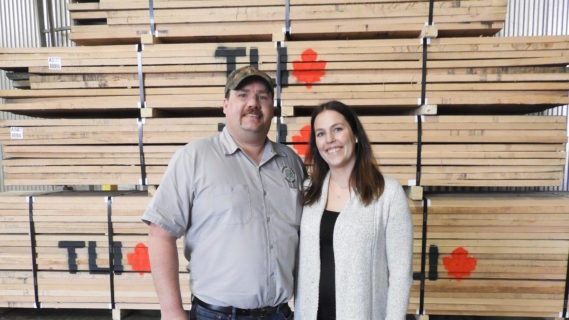 Man and women standing in front of a large pile of lumber with the TLI logo printed on the side.