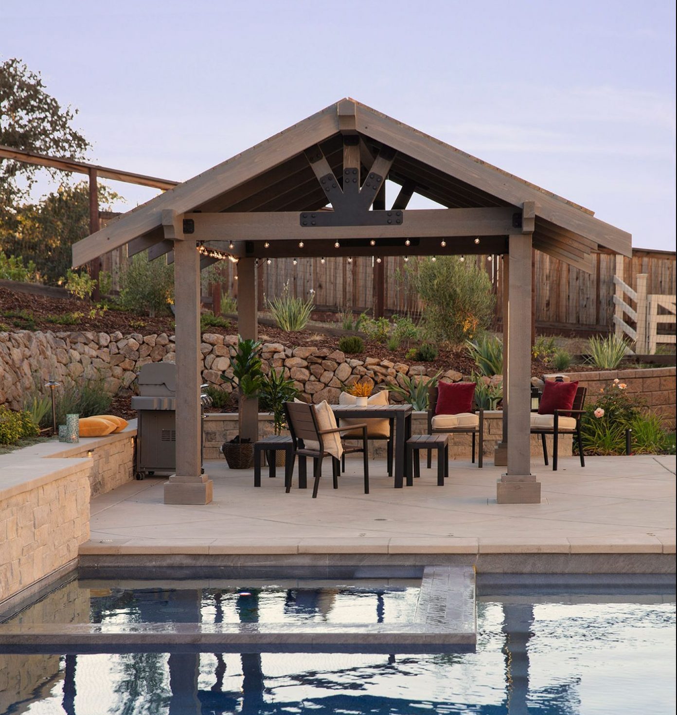 Wooden pergola with chairs and table next the swimming pool. Picture taken at dusk.