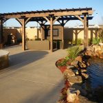 Outdoor Pergola covering a couch and tv on the patio next to a rocky pond.