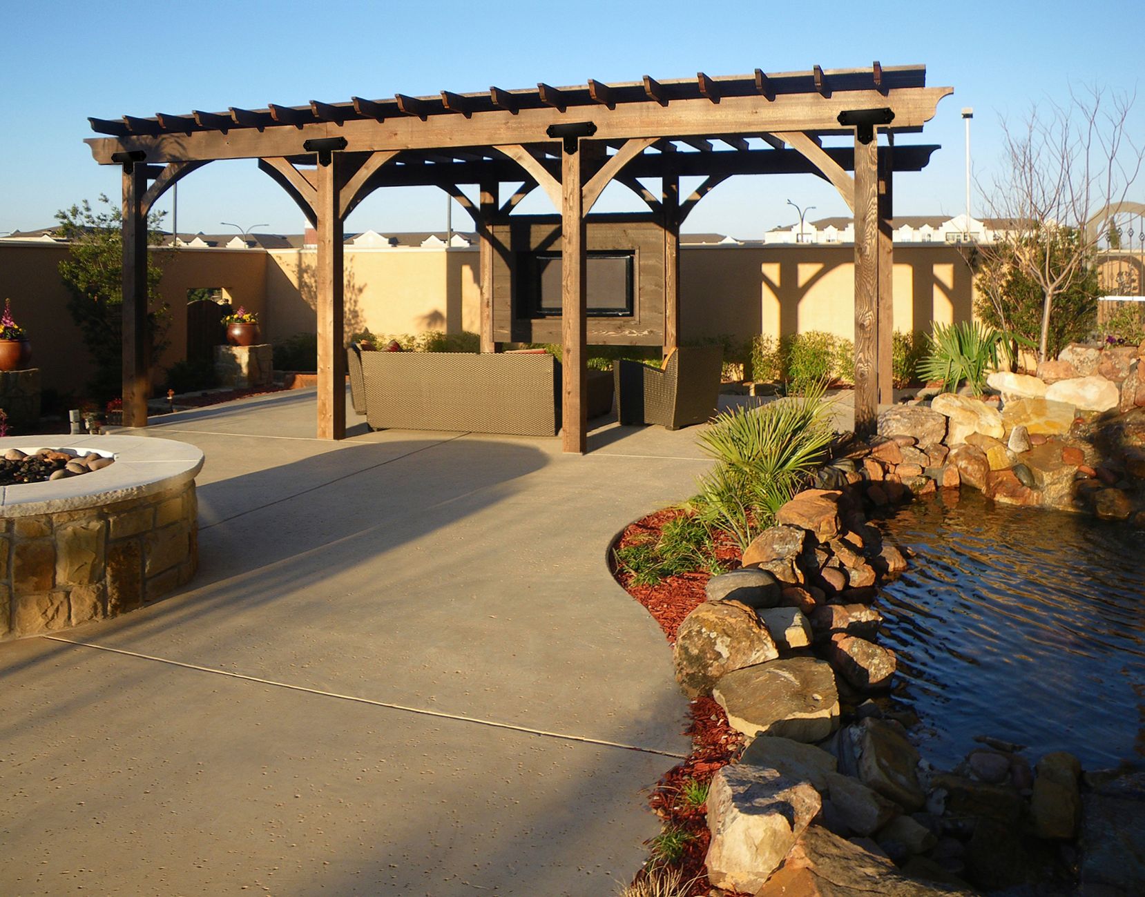 Outdoor Pergola covering a couch and tv on the patio next to a rocky pond.
