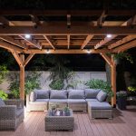 Pergola covering a wooden deck with couches and large pants. Photo taken at night.