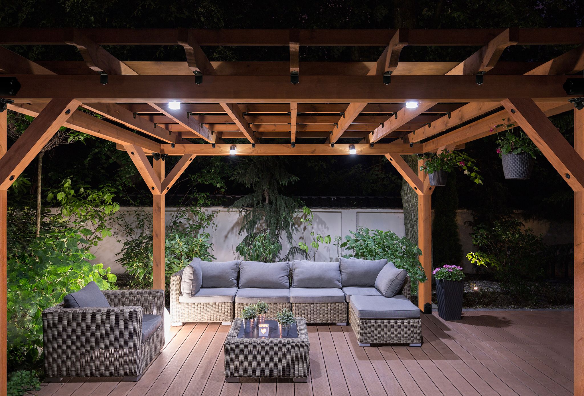 Pergola covering a wooden deck with couches and large pants. Photo taken at night.