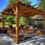 Wooden pergola with chairs and table next to a shed and large grass field. Picture taken on a sunny day.