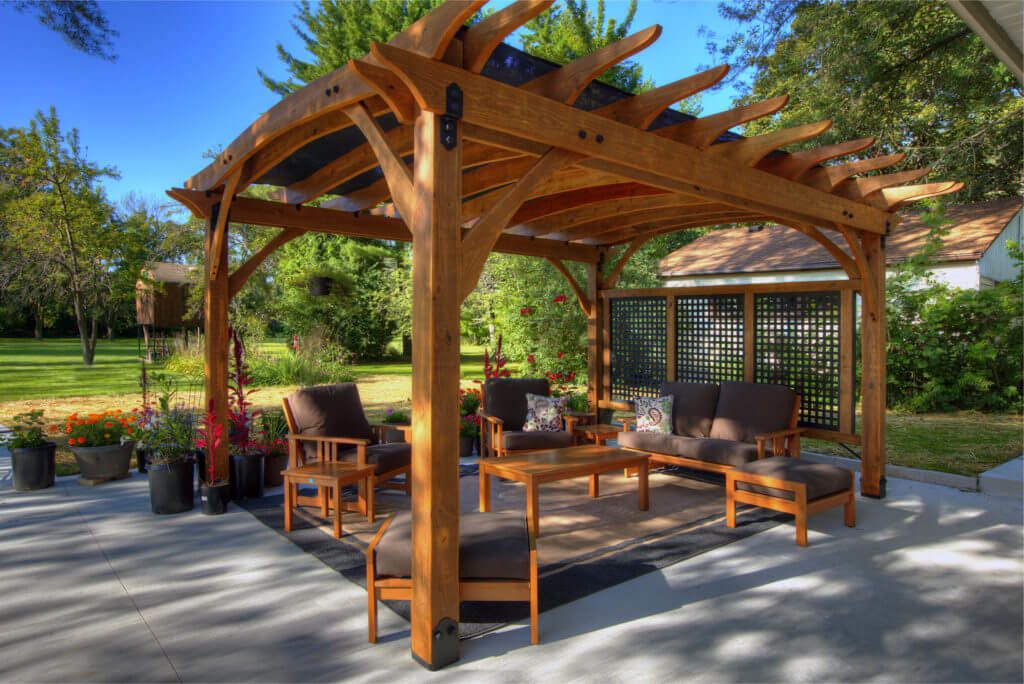 Wooden pergola with chairs and table next to a shed and large grass field. Picture taken on a sunny day.